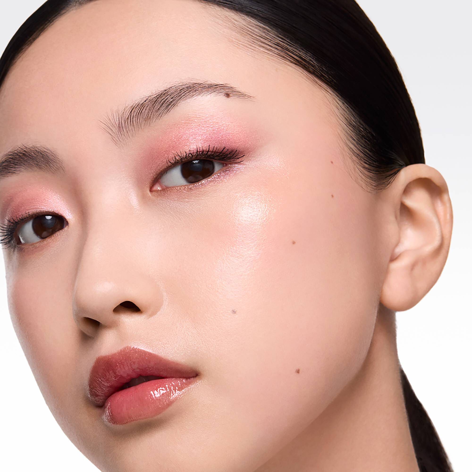 A close-up portrait of a woman with radiant skin, wearing soft pink shimmery eyeshadow and a glossy, berry-toned lip.	