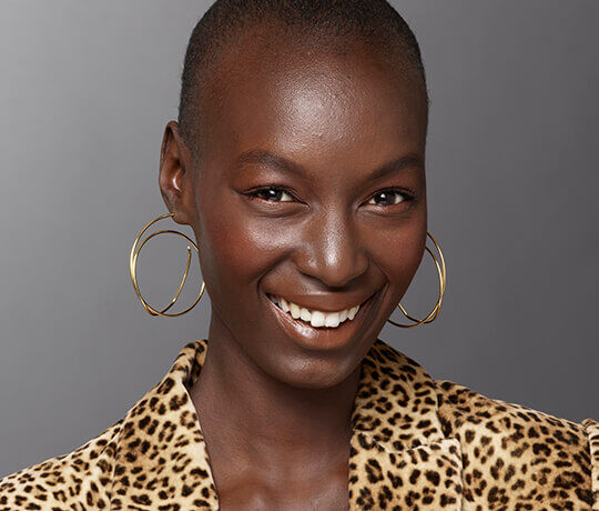 A smiling woman with a shaved head is wearing large gold hoop earrings and a leopard print shirt. She has a warm expression against a neutral background.