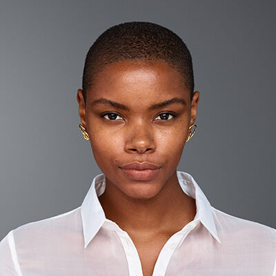 A close-up portrait of a young woman with short hair, wearing a white button-up shirt and gold earrings, against a gray background. She has a neutral expression and natural makeup.