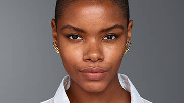 A close-up portrait of a young woman with short hair, wearing a white button-up shirt and gold earrings, against a gray background. She has a neutral expression and natural makeup.