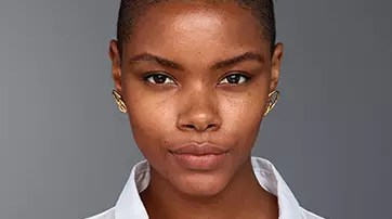 A close-up portrait of a young woman with short hair, wearing a white button-up shirt and gold earrings, against a gray background. She has a neutral expression and natural makeup.