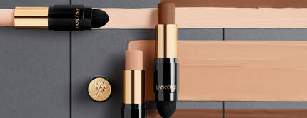 A close-up of Lancome makeup sticks in various shades displayed against a backdrop of swatches of foundation colors.
