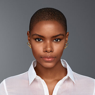 A close-up portrait of a confident young woman with short hair, wearing a white shirt and distinctive gold earrings, set against a neutral gray background.