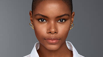 A close-up portrait of a confident young woman with short hair, wearing a white shirt and distinctive gold earrings, set against a neutral gray background.