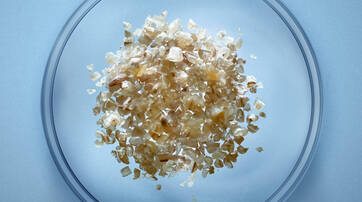 A glass petri dish containing a mound of light brown and beige sea salt crystals, set against a smooth blue background.