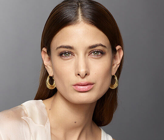 A close-up portrait of a model with long, straight hair and green eyes, wearing gold hoop earrings. She has a natural makeup look, with a subtle lip color, and is posed against a soft grey background.