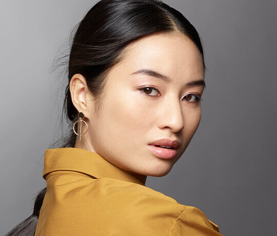 A close-up portrait of a young woman with long, dark hair, wearing a mustard-colored shirt and unique earrings. She gazes over her shoulder with a soft expression against a neutral background.