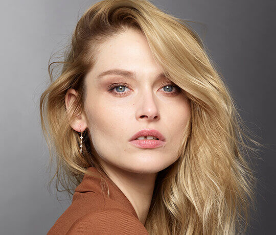 Close-up portrait of a woman with wavy blonde hair and blue eyes, wearing subtle makeup and silver earrings, against a soft gray background.