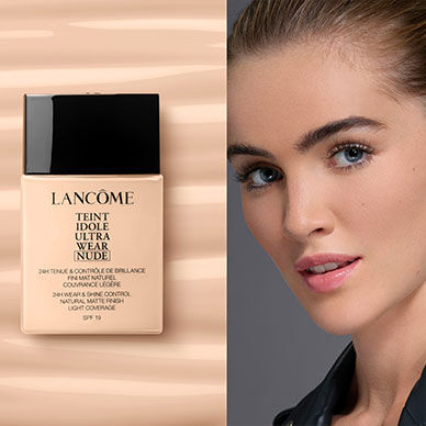 A close-up of a model's face next to a bottle of Lancome Teint Idole Ultra Wear Nude foundation, set against a neutral background with fluid beige tones.