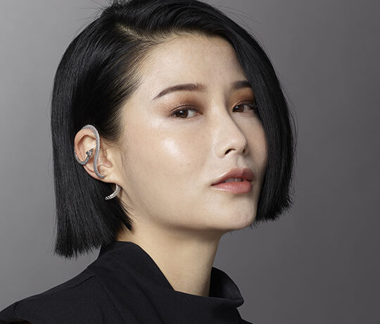 A close-up portrait of a woman with short black hair, wearing a stylish outfit and a unique silver ear accessory. She has a subtle makeup look and is posed against a neutral background.
