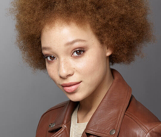 A close-up portrait of a young woman with curly brown hair and freckles, wearing a brown leather jacket. She has a gentle expression and is looking directly at the camera against a neutral background.