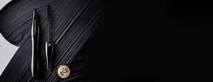 A sleek black mascara tube and wand are elegantly placed on a textured black background, with a gold logo visible nearby.