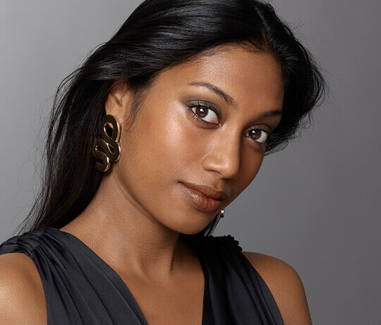 A woman with long, straight black hair is looking towards the camera, wearing bold, gold earrings and a dark top. Her makeup is natural, highlighting her features.