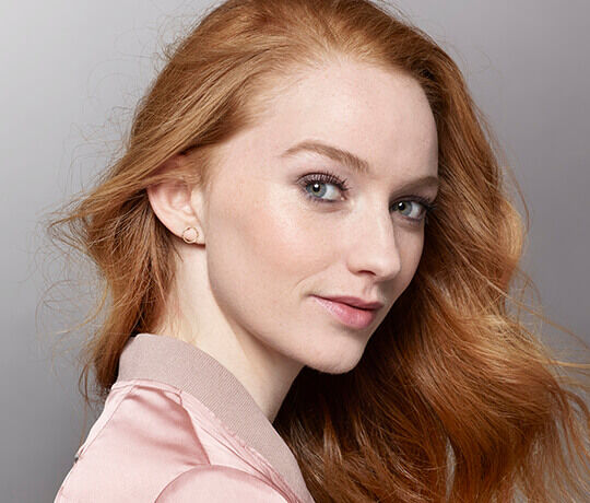 A young woman with long, reddish hair is smiling slightly, looking over her shoulder. She is wearing a light pink bomber jacket and has subtle makeup, highlighting her features. The background is softly blurred.