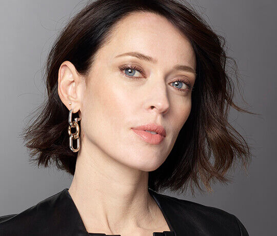 A woman with short, dark hair and a subtle makeup look poses confidently, wearing large, modern earrings and a black top.