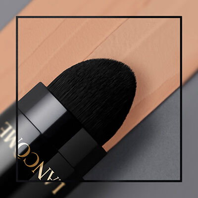 Close-up of a black makeup brush with a rounded tip, resting beside a swatch of peach-colored foundation on a smooth surface.