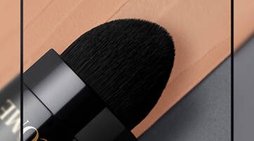 Close-up of a black makeup brush with a rounded tip, resting beside a swatch of peach-colored foundation on a smooth surface.