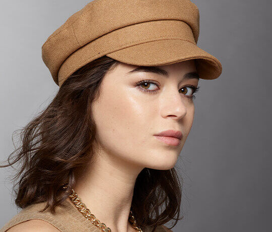 A young woman with wavy hair wearing a tan cap and a gold chain necklace, gazing slightly to the side against a gray background.