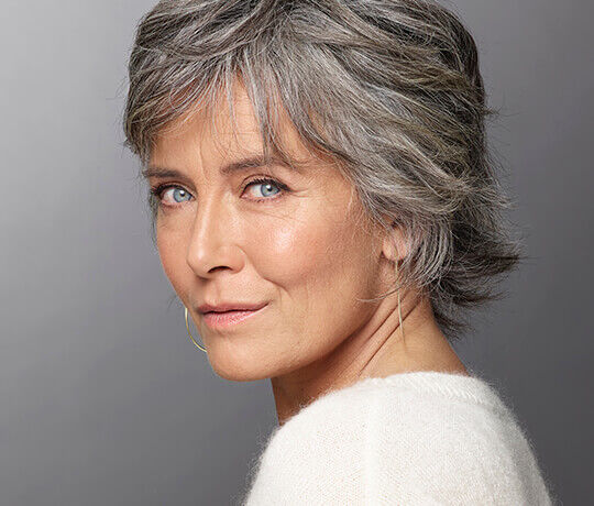 A woman with short, gray hair and blue eyes is looking over her shoulder, wearing a light sweater. She has a subtle smile and is posed against a soft, neutral background.