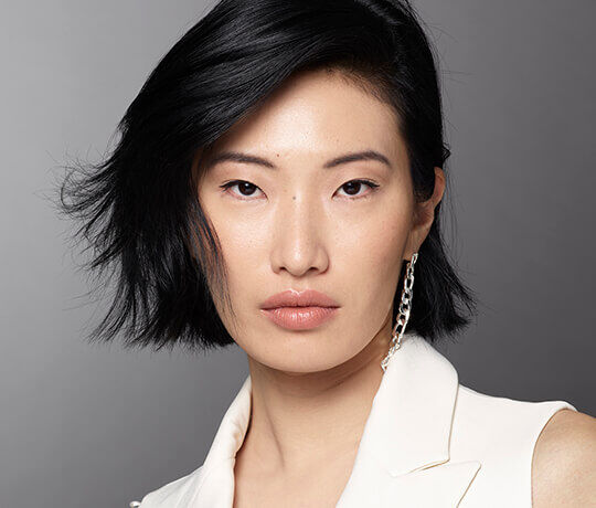 A close-up portrait of a person with black, shoulder-length hair and fair skin, wearing a white blazer. Their expression is neutral, and they have subtle makeup. A silver chain earring is visible on one ear, and the background is a soft gray gradient.