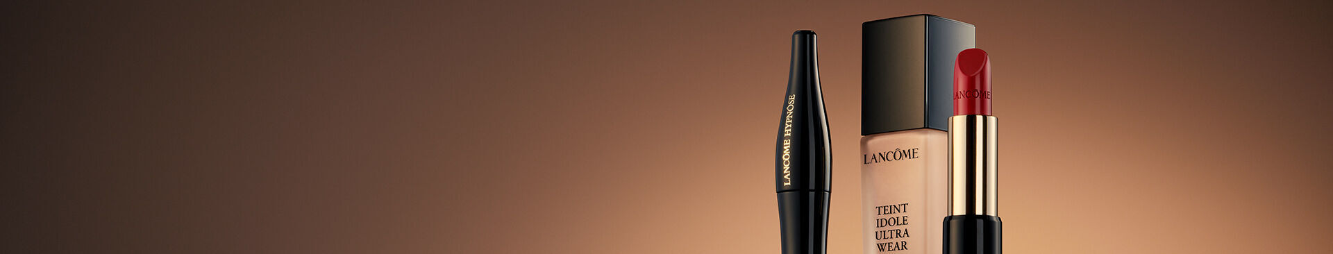 A close-up image of three makeup products against a warm, gradient background: a black eyeliner pencil, a foundation bottle, and a red lipstick.