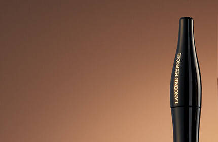 A close-up image of three makeup products against a warm, gradient background: a black eyeliner pencil, a foundation bottle, and a red lipstick.