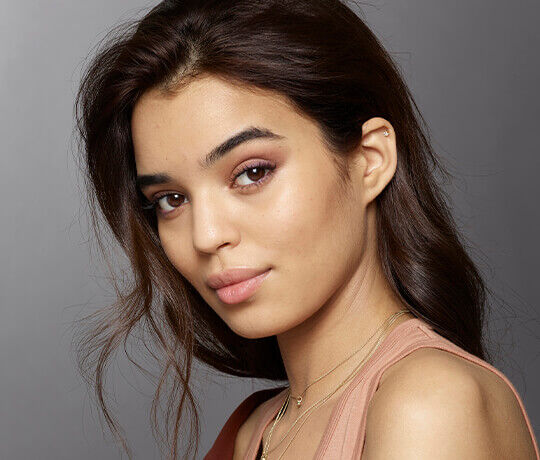 A close-up portrait of a young woman with long, wavy dark hair, wearing a soft pink top. She has a natural makeup look with a subtle eye shadow and a gentle smile. The background is a soft gray, enhancing her features.