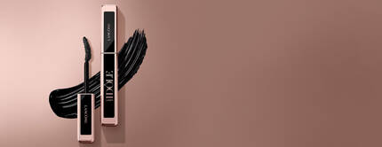 Two elegant mascara tubes from Lancome are displayed against a soft brown background, with a bold, sweeping stroke of black mascara accentuating the composition.