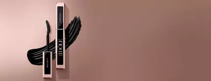 Two elegant mascara tubes from Lancome are displayed against a soft brown background, with a bold, sweeping stroke of black mascara accentuating the composition.