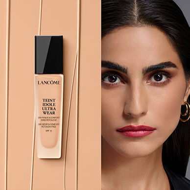 A sleek bottle of Lancome Teint Idole Ultra Wear foundation stands against a beige background, next to a close-up of a woman with a flawless complexion, bold eyebrows, and striking red lips.