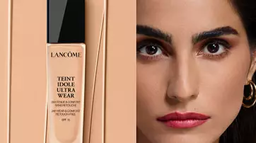 A sleek bottle of Lancome Teint Idole Ultra Wear foundation stands against a beige background, next to a close-up of a woman with a flawless complexion, bold eyebrows, and striking red lips.