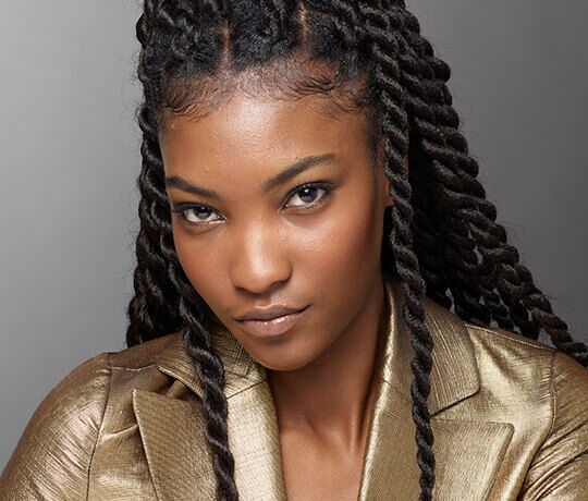 A close-up portrait of a person with long, twisted hair, wearing a shiny gold jacket, looking directly at the camera with a confident expression.