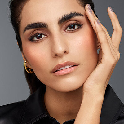 A close-up of a young woman with dark brows and long lashes, touching her face gently. She has a natural makeup look, with a subtle blush and nude lips, wearing a black leather top and hoop earrings. The background is a soft gray.