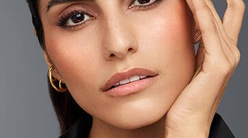 A close-up of a young woman with dark brows and long lashes, touching her face gently. She has a natural makeup look, with a subtle blush and nude lips, wearing a black leather top and hoop earrings. The background is a soft gray.