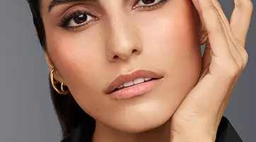 A close-up of a young woman with dark brows and long lashes, touching her face gently. She has a natural makeup look, with a subtle blush and nude lips, wearing a black leather top and hoop earrings. The background is a soft gray.