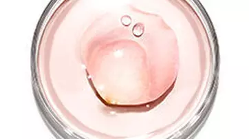 A close-up view of a clear jar containing a pale pink gel with a droplet of liquid resting on top.