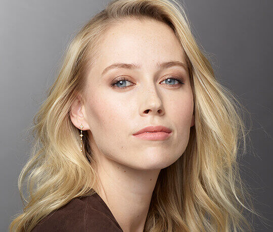 A close-up portrait of a woman with blonde, wavy hair, soft makeup, and wearing earrings, gazing directly at the camera.