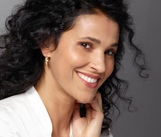 A woman with curly hair smiles warmly, wearing gold hoop earrings and a white top. She rests her chin on her hand against a neutral gray background.