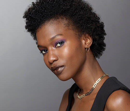 A close-up portrait of a model with short, curly hair, wearing bold purple eyeshadow and glossy brown lips. She accessorizes with hoop earrings and a gold chain necklace, and is looking confidently over her shoulder against a neutral gray background.