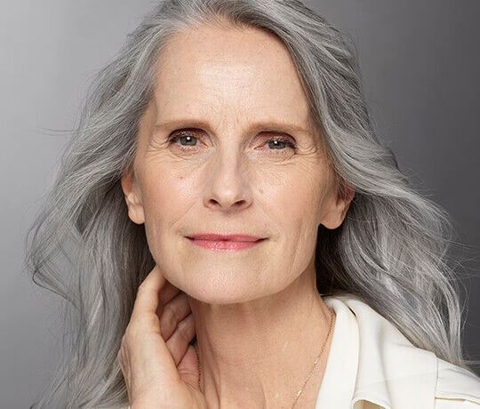 A mature woman with long, gray hair smiles softly, showing natural skin texture and a calm expression. She is wearing a light-colored blouse and gently touches her neck with one hand.