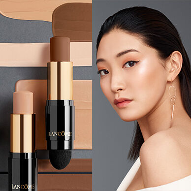 A close-up image featuring two sticks of Lancome foundation alongside varying shades of foundation swatches. On the right, a model with long dark hair and minimal makeup poses, showcasing a sleek look.