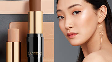 A close-up image featuring two sticks of Lancome foundation alongside varying shades of foundation swatches. On the right, a model with long dark hair and minimal makeup poses, showcasing a sleek look.