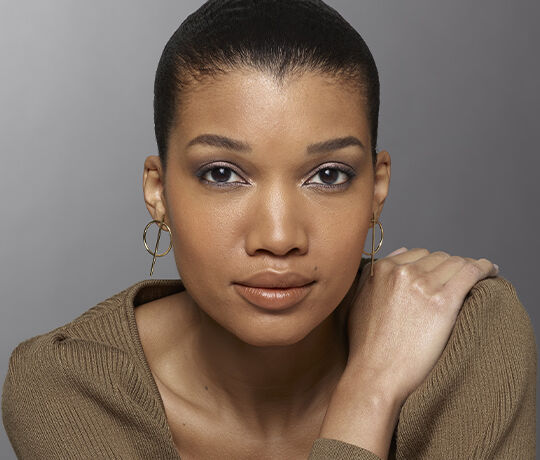 A close-up portrait of a woman with a smooth complexion, wearing elegant gold hoop earrings. She has light makeup with a focus on her eyes, and her hair is styled in a sleek bun. She is looking directly at the camera with a neutral expression, dressed in a soft, tan sweater. The background is a gradient gray.