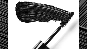 A close-up of a mascara wand with a bold black brushstroke beside it, set against a white background.