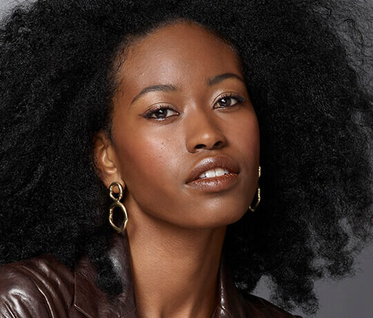 Close-up portrait of a woman with natural, voluminous black hair, wearing gold hoop earrings and a brown leather jacket. She has a natural makeup look, highlighting her features.