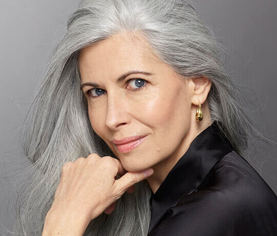 A close-up portrait of a woman with long gray hair, fair skin, and a slight smile, resting her chin on her hand while wearing a black top and gold earrings.