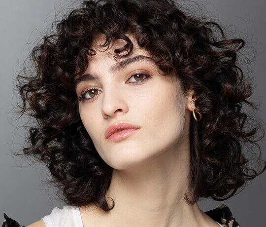 A young woman with curly hair and soft makeup gazes confidently at the camera, wearing a simple white top and a gold hoop earring.
