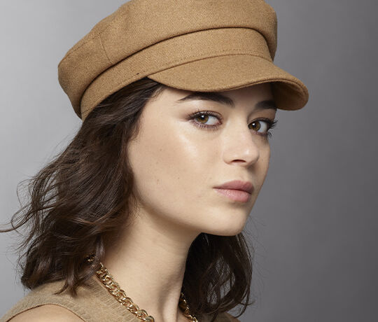 A young woman with long, dark hair is turned slightly to the side, wearing a tan cap and a gold chain necklace. She has a neutral expression against a gray background.