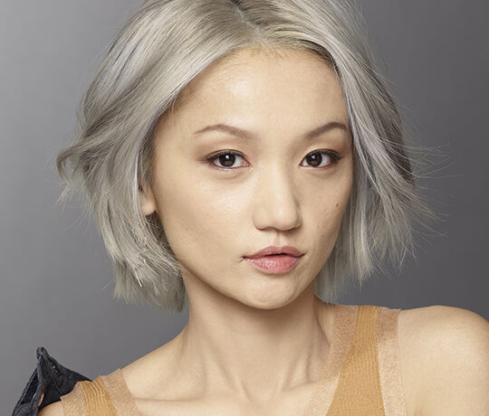 A close-up portrait of a person with short, textured silver hair and natural makeup, looking directly at the camera with a neutral expression.