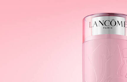 Two Lancome skincare products are displayed against a soft pink background. The left product has a light pink cylindrical bottle with a silver cap, while the right product features a sleek design with a shiny silver lid.